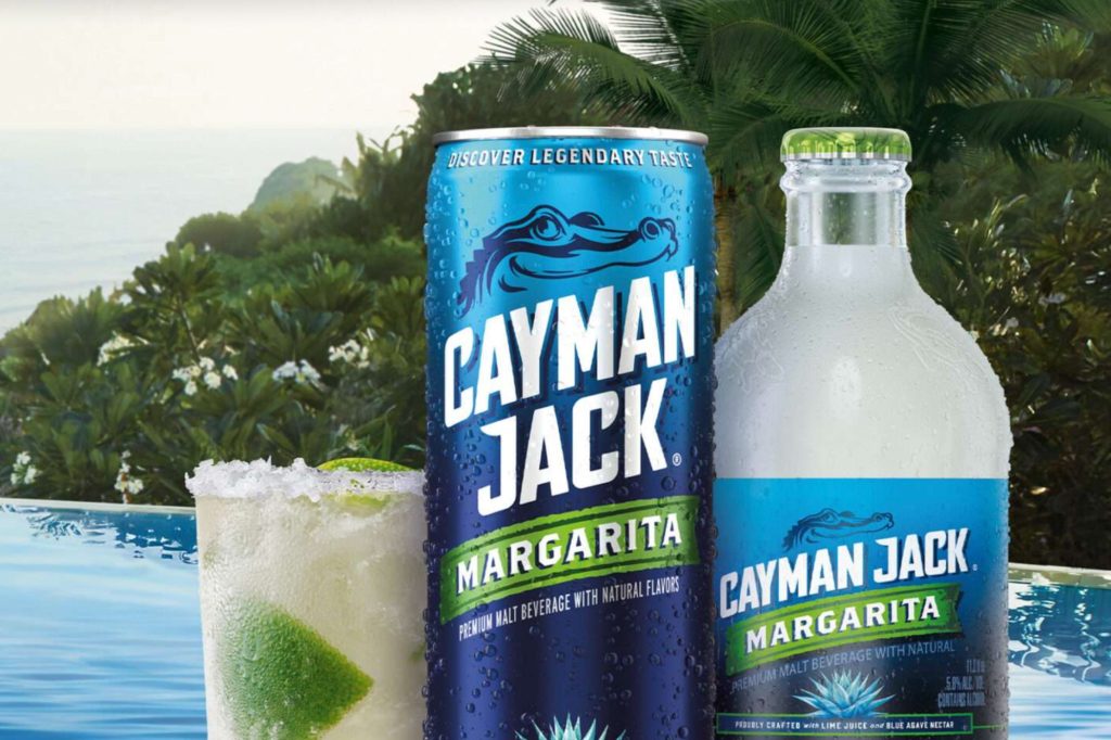 A refreshing margarita sits in a glass with lime and ice, accompanied by a can and bottle of a premium malt beverage, set against a scenic tropical backdrop, featured in the Cayman Jack Escape to Paradise Sweepstakes