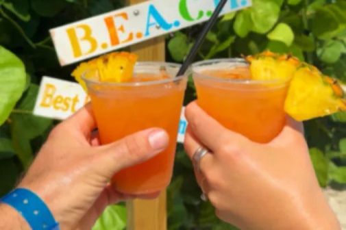 Two hands raise tropical drinks with pineapple garnishes in front of a colorful "B.E.A.C.H." sign, suggesting a cheerful beachside atmosphere, featured in the National Seashell Day Sweepstakes