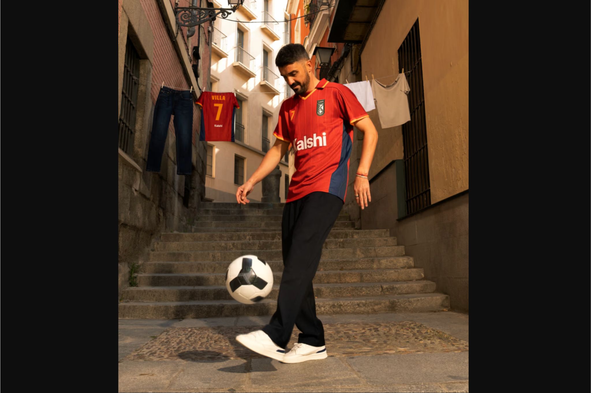 A man in a red jersey skillfully juggles a soccer ball while standing on steps. Clothes are hanging on a line nearby, and the scene captures a vibrant urban atmosphere, featured in the Kalshi $1,000,000 World Cup Sweepstakes