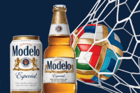 A can and a bottle of Modelo beer are positioned beside a colorful soccer ball hitting the back of a net, against a dark blue background, featured in the MODELO SUMMER SOCCER FAN EXPERIENCE SWEEPSTAKES