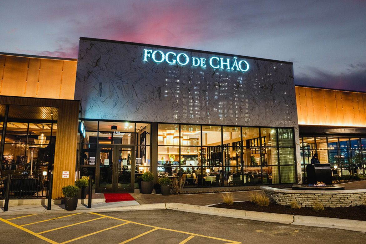 The modern exterior of a restaurant features large glass windows reflecting evening lights, a welcoming red carpet at the entrance, and a stylish facade illuminated against a dusky sky, featured in the Fogooooal Experience Sweepstakes