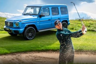 A golfer swings in a sand trap on a lush green course, with a stylish blue SUV parked nearby against a backdrop of rolling hills and a bright sky, featured in the Mercedes-Benz The Open Sweepstakes
