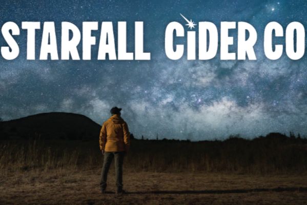 A person in a yellow jacket stands on a grassy field, gazing up at a stunning night sky filled with stars and the Milky Way, with a silhouette of a hill in the background, featured in the Created On Earth Sweepstakes