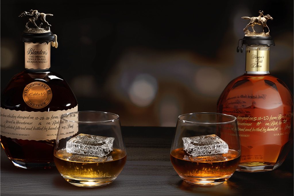 Two glasses of bourbon whiskey sit on a dark wooden surface, each containing a large ice cube, accompanied by two distinct bottles of bourbon, elegantly displayed, featured in the Eddie V’s Father’s Day Sweepstakes