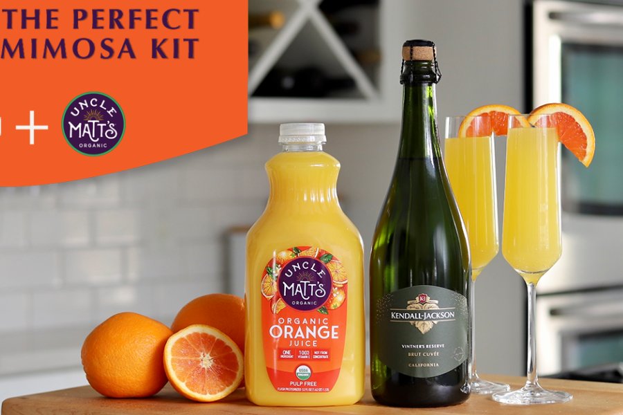 A bottle of organic orange juice and a bottle of sparkling wine sit on a table alongside three flutes of mimosas garnished with orange slices, creating a vibrant brunch setting, featured in the Kendall-Jackson Mimosas Sweepstakes
