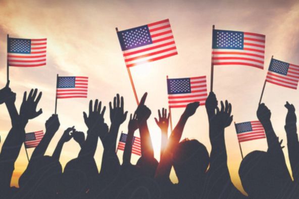 A crowd of people raises their hands and waves small American flags against a vibrant sunset, celebrating unity and patriotism, featured in the GOVX $10K Hero Giveaway