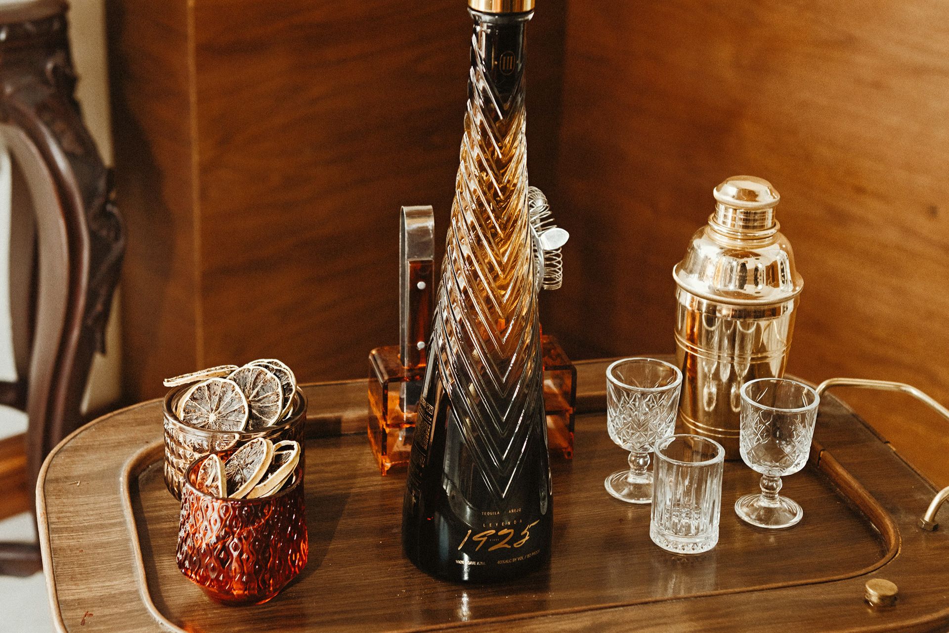 A luxurious bottle of tequila stands prominently on a wooden bar cart, surrounded by elegant glassware and decorative dried citrus slices in stylish containers, featured in the Leyenda 1925 Tickets Sweepstakes