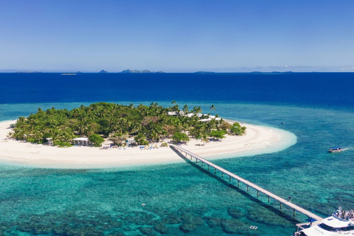 A tropical island with lush palm trees and a sandy beach is surrounded by clear turquoise waters, featuring a wooden pier extending from the shore toward the open sea, featured in the Fiji Airways Fiji Escape Sweepstakes