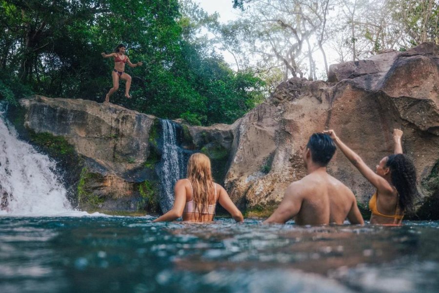 A person jumps off a rocky ledge into a natural pool, while three friends cheer from the water below, surrounded by lush greenery and the sound of a cascading waterfall, featured in the Get Outside In Costa Rica Giveaway