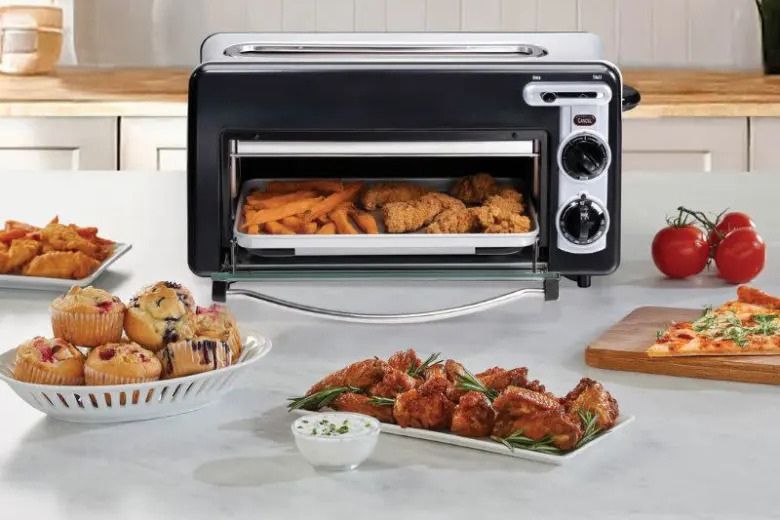 A countertop toaster oven displays a tray with chicken and French fries, surrounded by plates of muffins, chicken wings, and pizza, set in a bright kitchen, featured in the Hamilton Beach Toastation 2-in-1 Toaster Sweepstakes