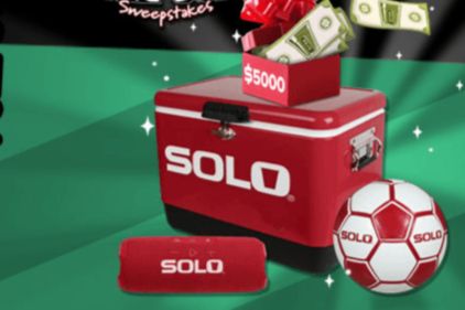 A red cooler sits on a green background, with a box of cash and a soccer ball nearby, creating a vibrant scene that suggests a fun and exciting giveaway or promotion, featured in the Solo Home Stadium Makeover Sweepstakes