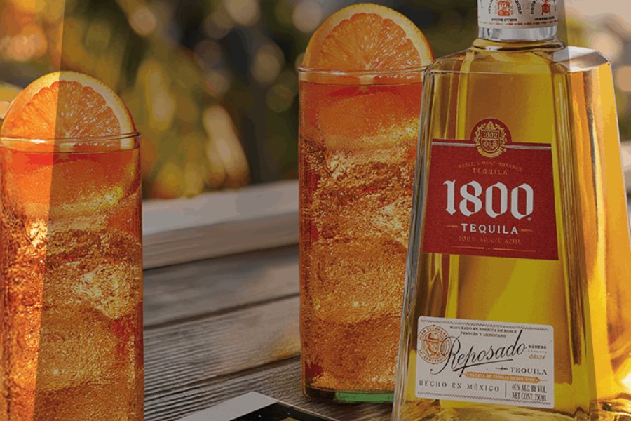 Two tall glasses filled with a sparkling orange drink, garnished with orange slices, sit next to an open bottle of tequila on a wooden surface in a bright setting, featured in the 1800 Tequila Elevate Your Summer Sweepstakes
