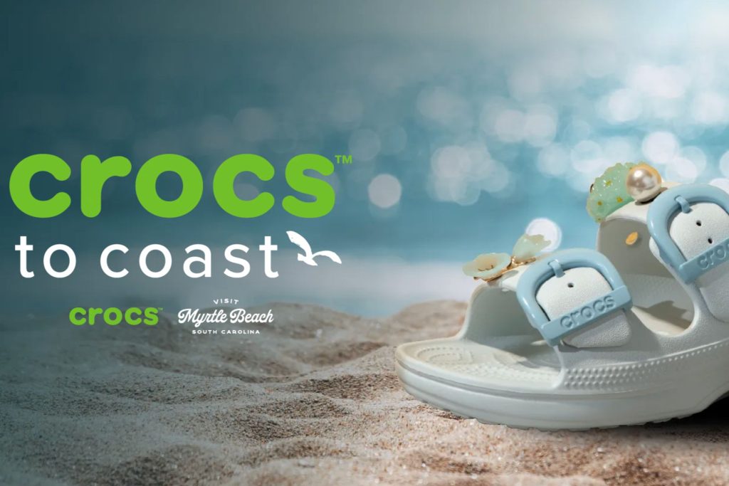 A pair of stylish white sandals with decorative accessories rests on soft beach sand, with blurred blue water and sparkling sunlight in the background, evoking a coastal vibe, featured in the Crocs to Coast Sweepstakes