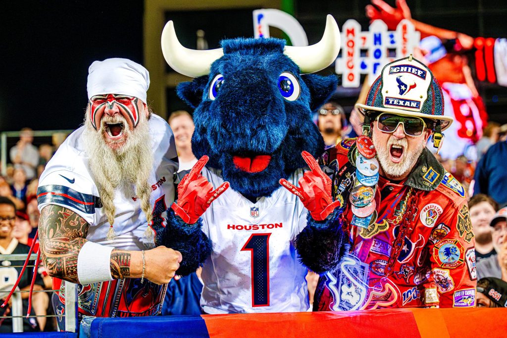 Excited fans in colorful attire pose together, with one fan in a Houston jersey, a mascot in blue and white, and another in a decorated outfit, celebrating at a lively sporting event, featured in the Genie Hall of Fame Trip Sweepstakes